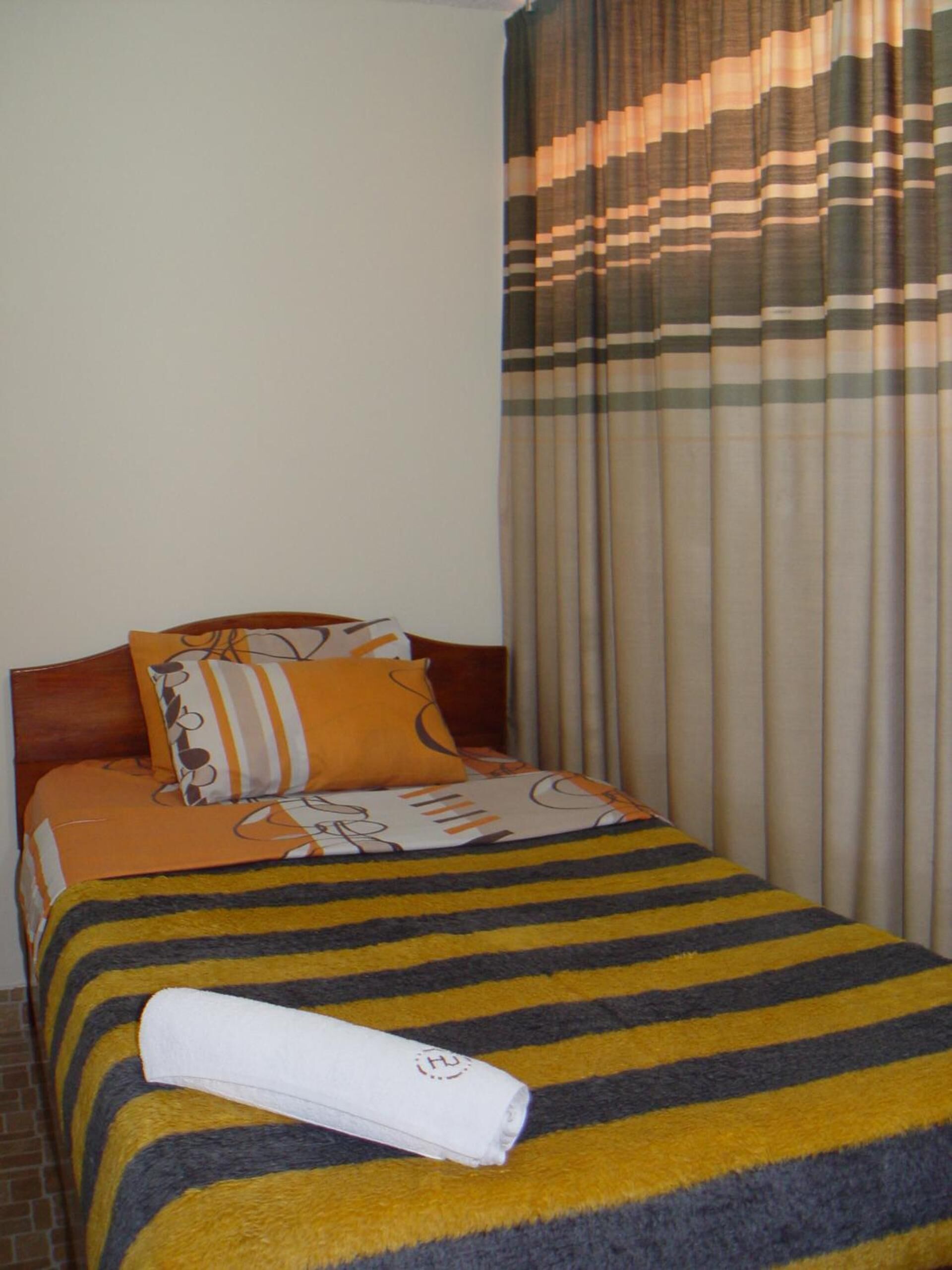 A hotel room with a bed, a wooden headboard, striped bedding, and a rolled towel on the bed.