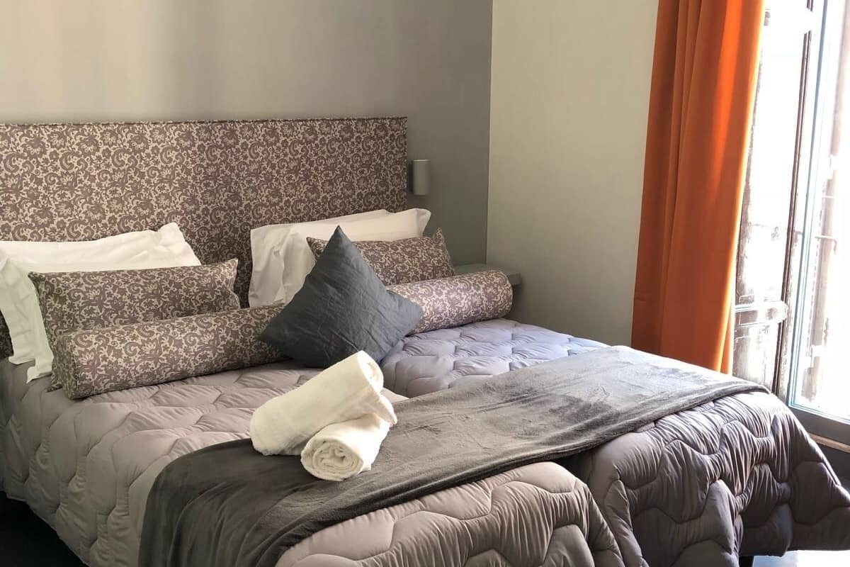 Una cama bien tendida con un cabecero estampado, una lámpara de noche y una ventana con cortinas.