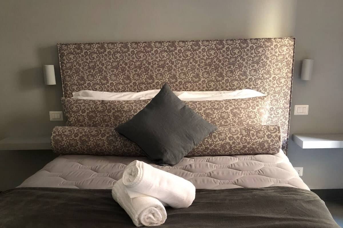 Una cama bien tendida con un cabecero estampado, una almohada oscura y toallas enrolladas sobre la cama.