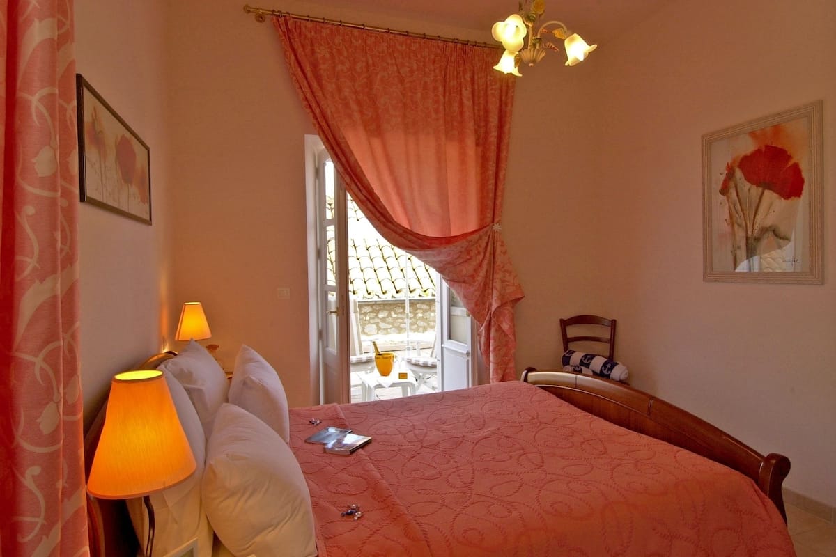 A bedroom with a bed, a nightstand, a lamp, a chair, a window with curtains, a painting, and a chandelier.