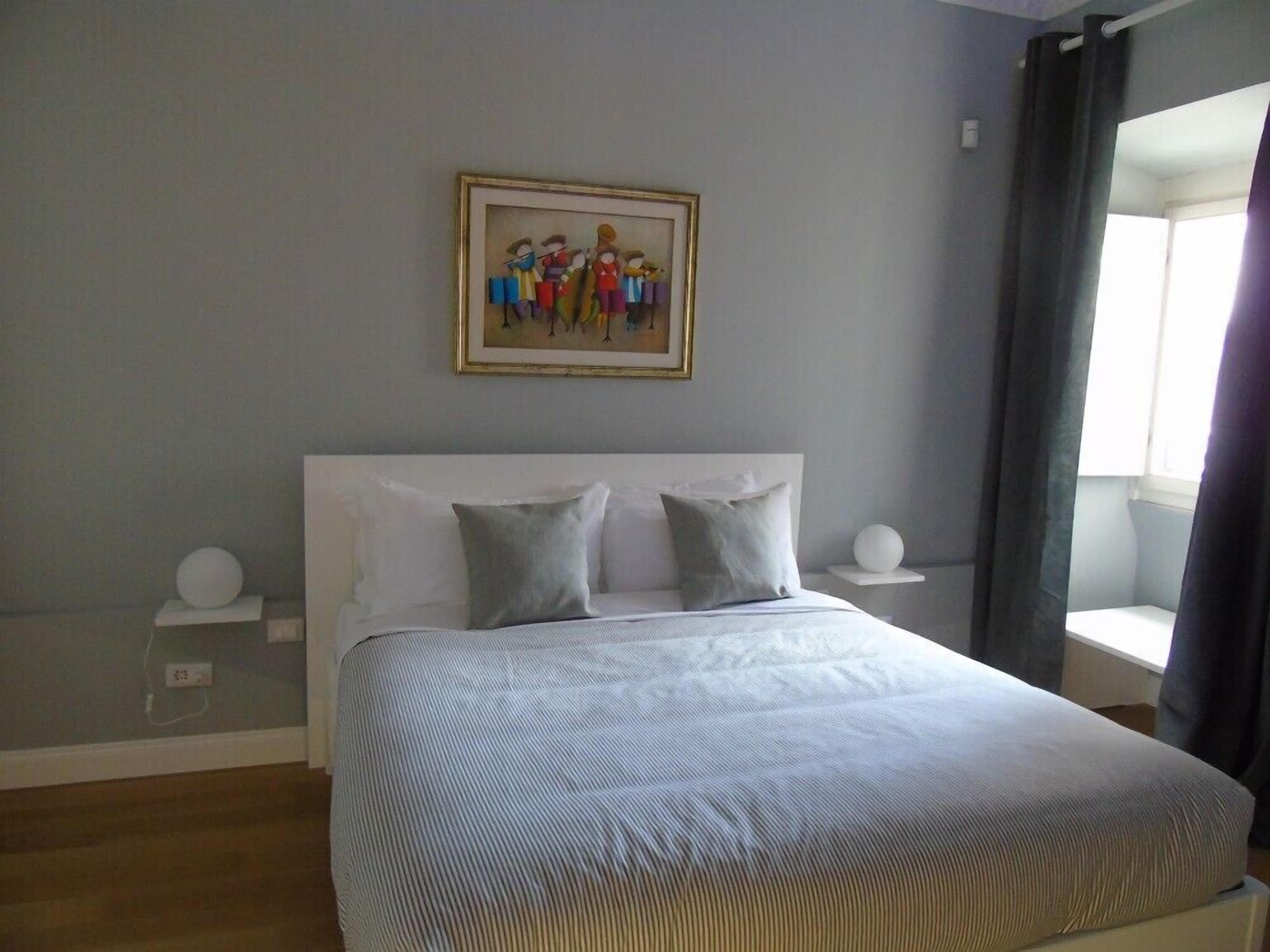 A bedroom with a large bed, a headboard, two bedside tables, a wall-mounted painting, and a window with curtains.