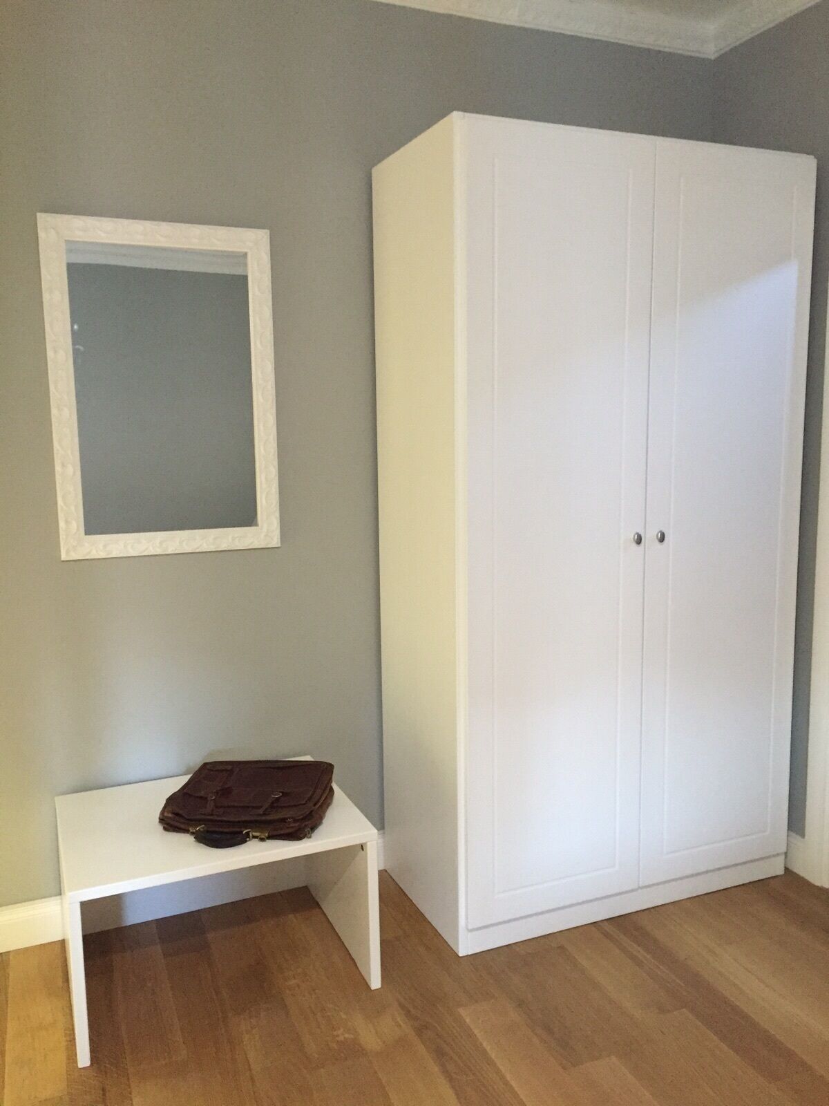 A room with a white wardrobe, a small white bench, and a framed mirror on the wall.