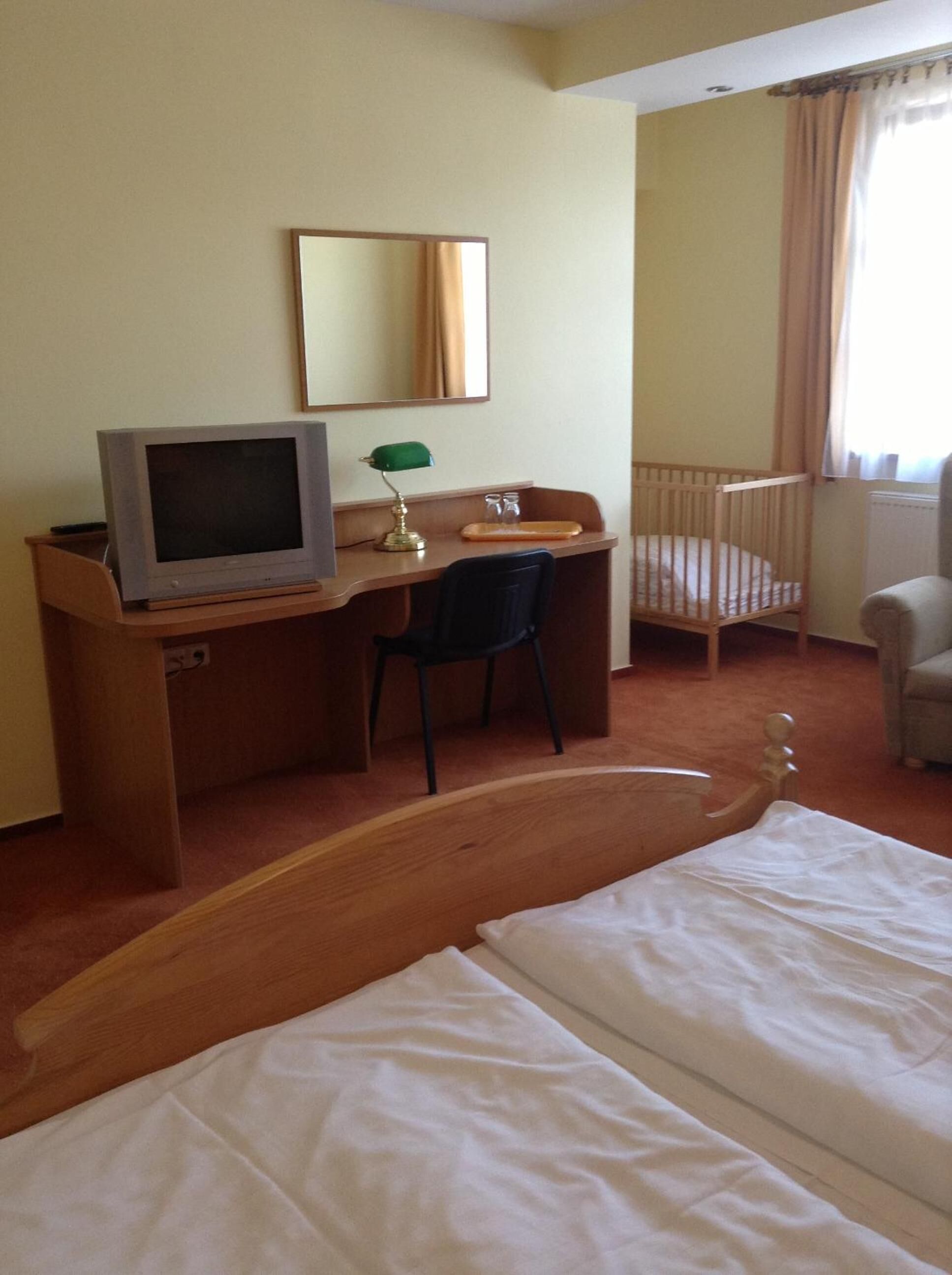 A hotel room with a bed, a desk with a television, a chair, a crib, and a window with curtains.