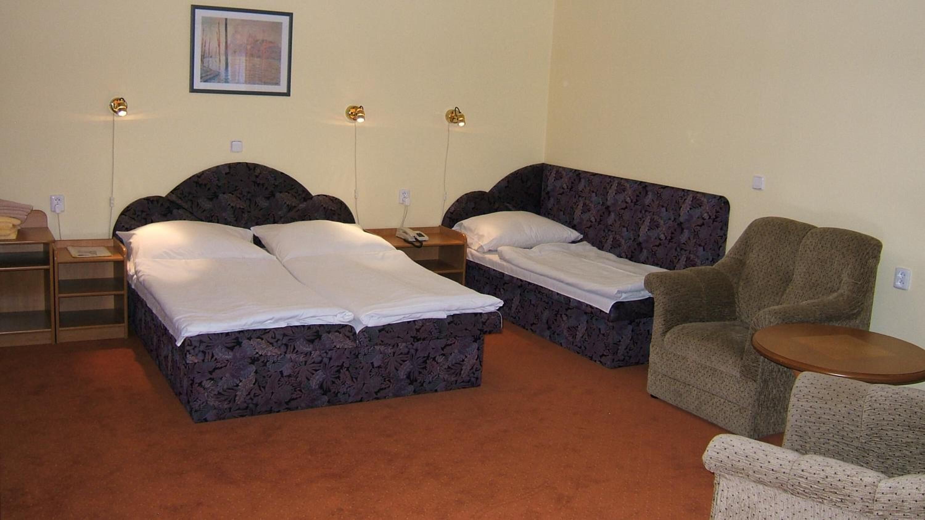 A room with two beds, a chair, a small table, and a framed picture on the wall.