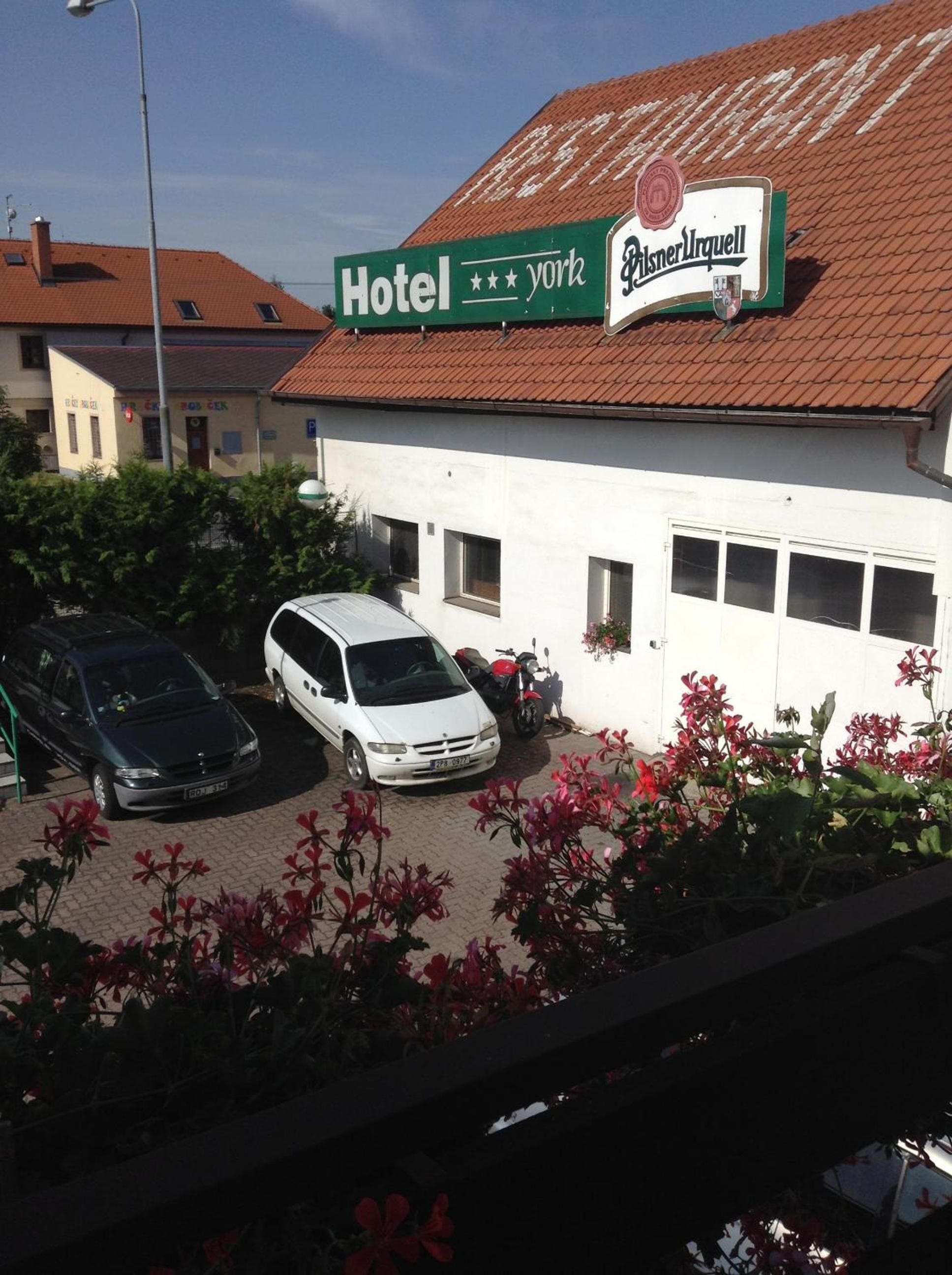A building with a sign that reads "Hotel York" and "Pilsner Urquell" on the roof. There are parked cars in front and a garden with red flowers in the foreground.