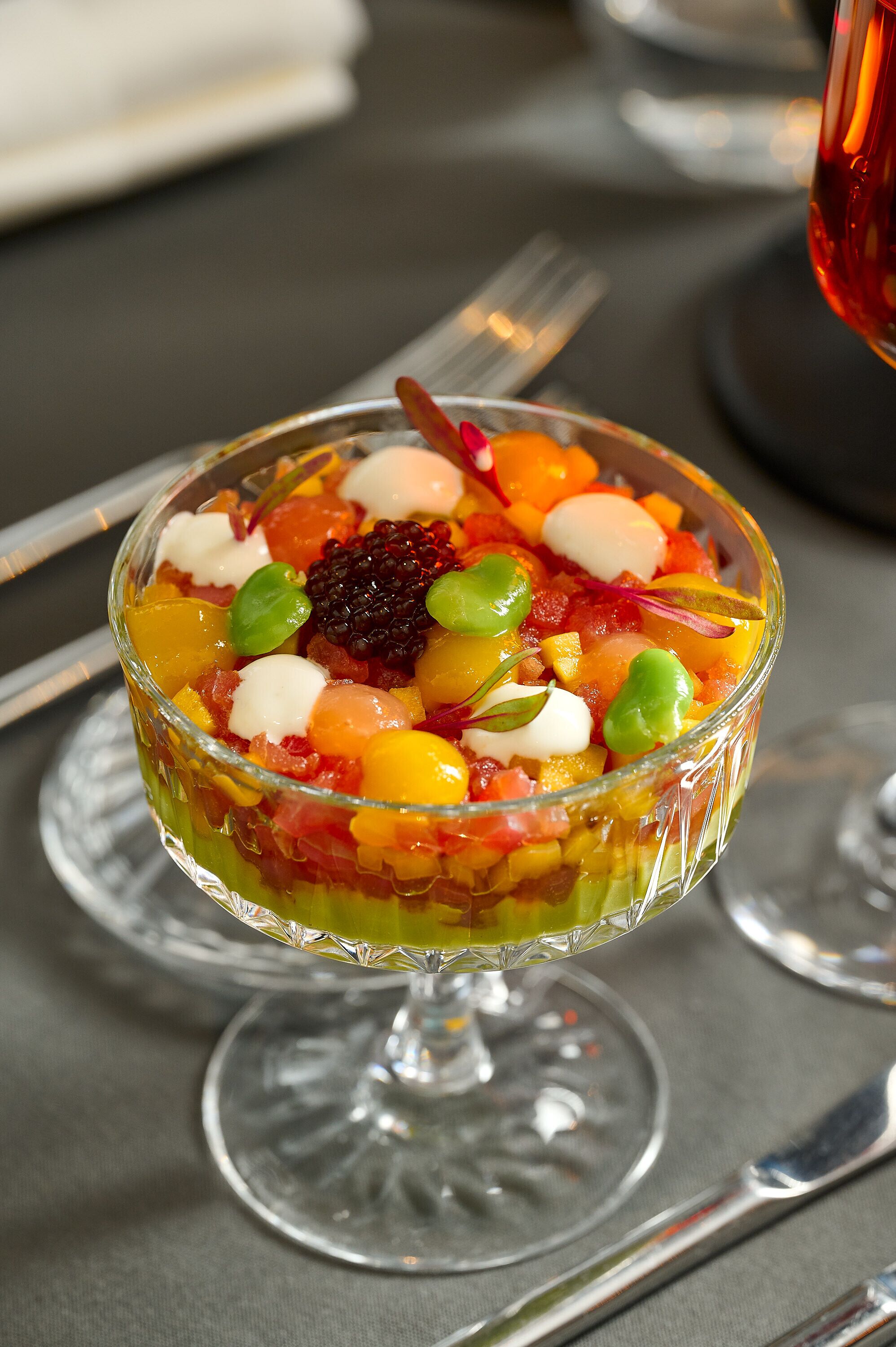 A clear glass dessert bowl on a table with a fork and a wine glass nearby.
