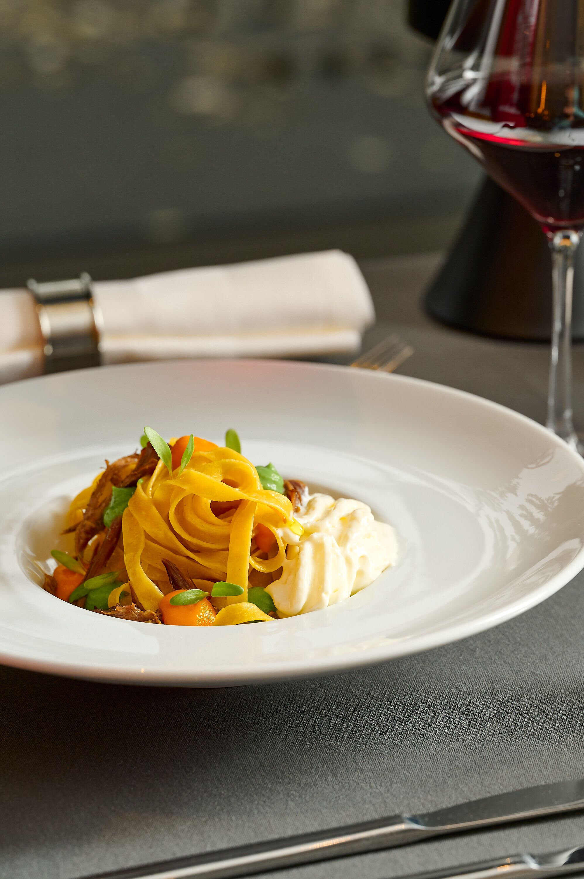 A white plate with a dish of pasta and vegetables, a wine glass, and a fork on a table.