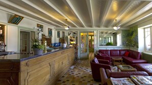 A spacious lobby with a checkered floor, a wooden bar, red leather seating, and a large window with a view of the outdoors. - Hôtel le Pré Serroux (Champagney)