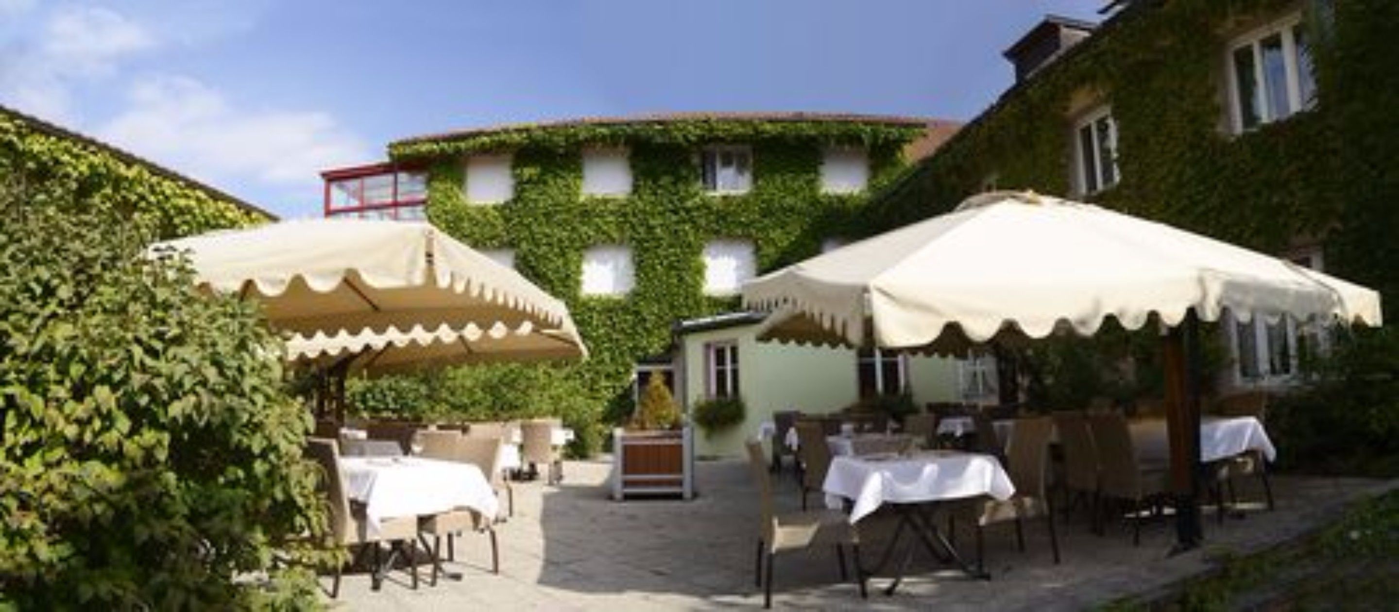 An outdoor patio area with tables and chairs, covered by white umbrellas, and surrounded by greenery.