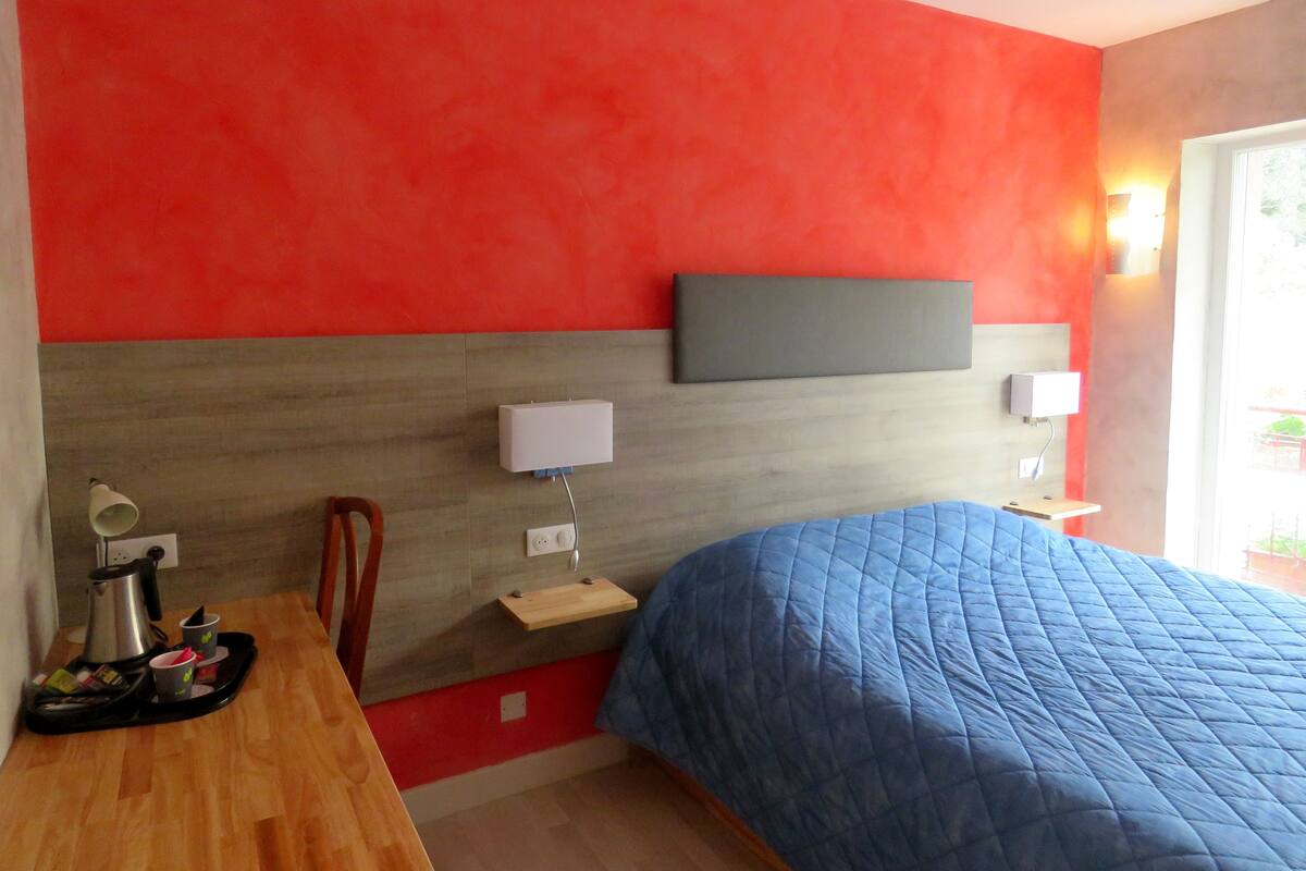 A hotel room with a red and grey headboard, a blue bedspread, a wooden desk with a kettle, cups, and a tray, and a small shelf with a lamp and a power outlet.