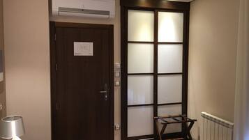 Habitación de hotel con una puerta de madera oscura, una puerta corredera de cristal con paneles satinados, una cama con edredón blanco, una lámpara de noche y un pequeño banco de madera cerca de la puerta.