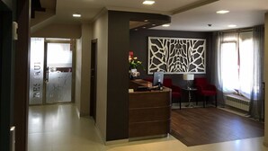 Un espace d’accueil d’hôtel au design moderne, comprenant un comptoir de réception, deux fauteuils rouges, un panneau mural décoratif et une fenêtre avec des rideaux.