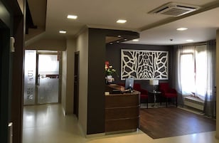 Un área de recepción de hotel con diseño moderno, que incluye un mostrador de recepción, dos sillas rojas, un panel decorativo en la pared y una ventana con cortinas.