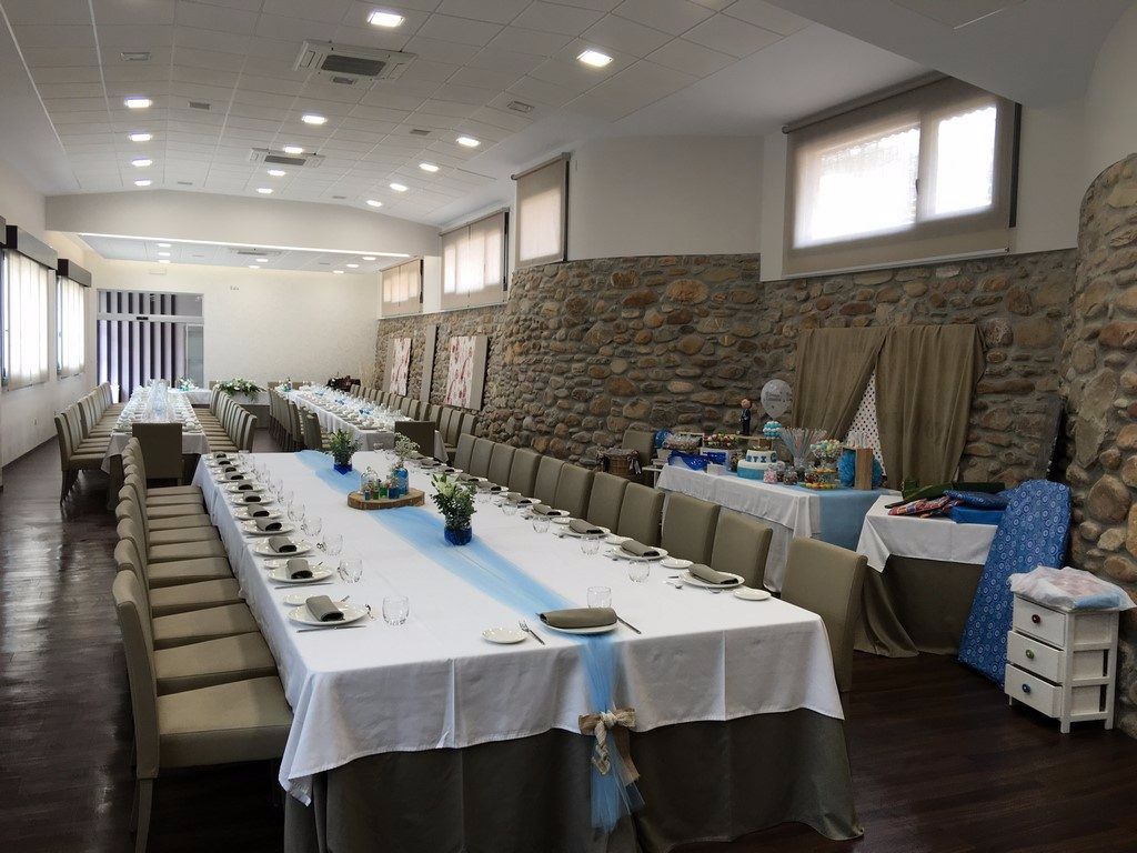 A long dining table set for a formal event, with chairs on both sides, in a room with stone walls and large windows.