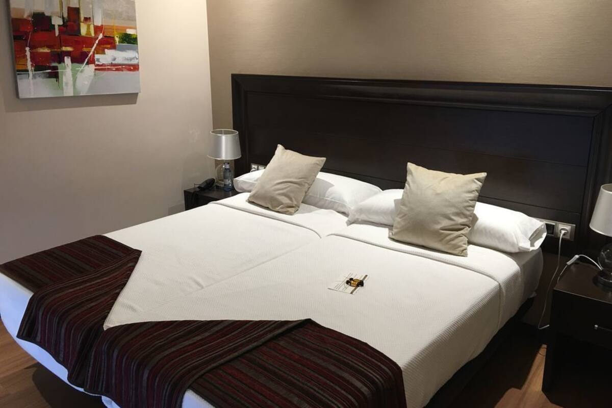 A neatly made bed with a white comforter and two pillows, a striped throw blanket, a bedside table with a lamp, and a wall-mounted abstract painting.