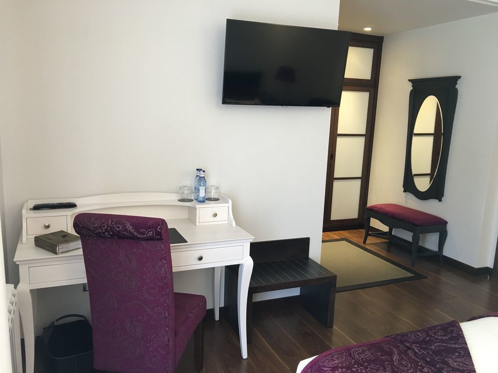 Une chambre d’hôtel avec un bureau blanc, une chaise violette, un téléviseur à écran plat fixé au mur, un petit banc et un miroir.