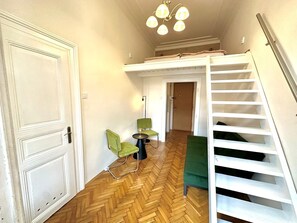A room with a herringbone wooden floor, a white door, a green chair, a small round table, and a white staircase with a green cushion.