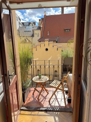 A balcony with a small table and chair, a potted plant, and a view of buildings with red-tiled roofs.