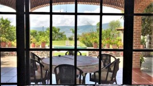 A patio area with a round table and chairs, a cushioned bench, and a view of a landscaped garden and mountains through large windows. - Villa Meli 2 by Kivoya (Jocotepec)