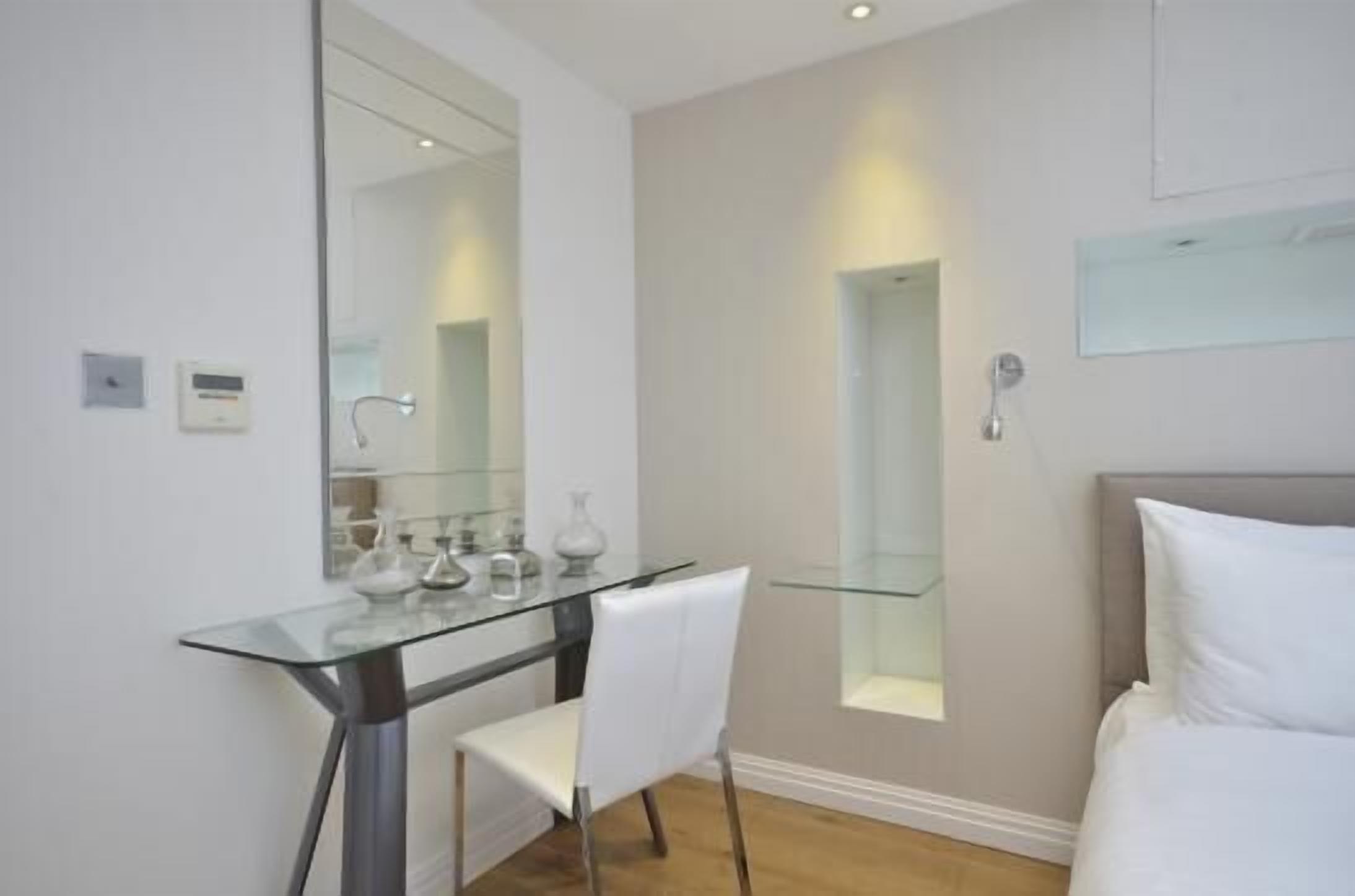 Een moderne hotelkamer met een glazen bureau, een witte stoel en een bed met witte beddengoed.