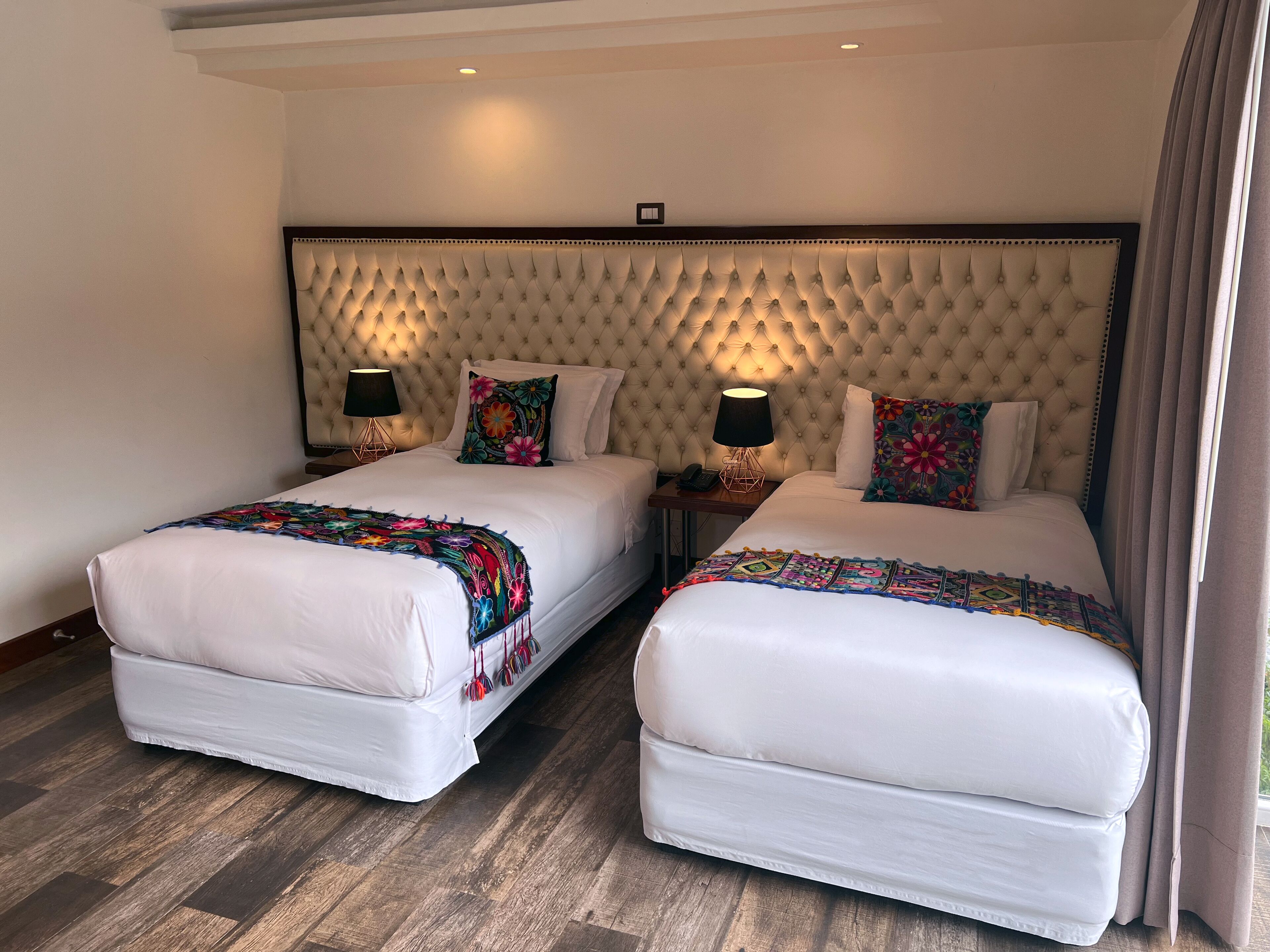 Quarto de hotel com duas camas, cada uma com cabeceira estofada, roupa de cama branca e almofadas decorativas florais coloridas. Há luminárias de cabeceira em mesas de cabeceira de madeira e o quarto possui piso de madeira.