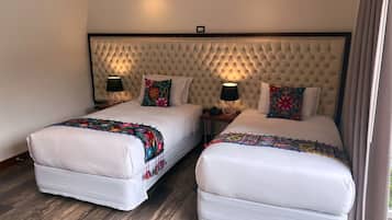 Quarto de hotel com duas camas, cada uma com cabeceira estofada, roupa de cama branca e almofadas decorativas florais coloridas. Há luminárias de cabeceira em mesas de cabeceira de madeira e o quarto possui piso de madeira.