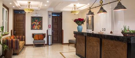 Una hall d'albergo con una reception, un divano con cuscini colorati, un cestino e una pianta in vaso.