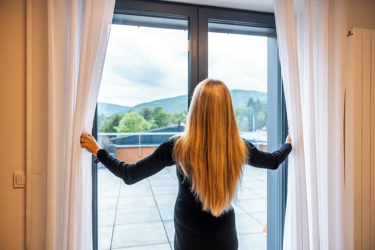 A person standing at a window with curtains, looking outside.