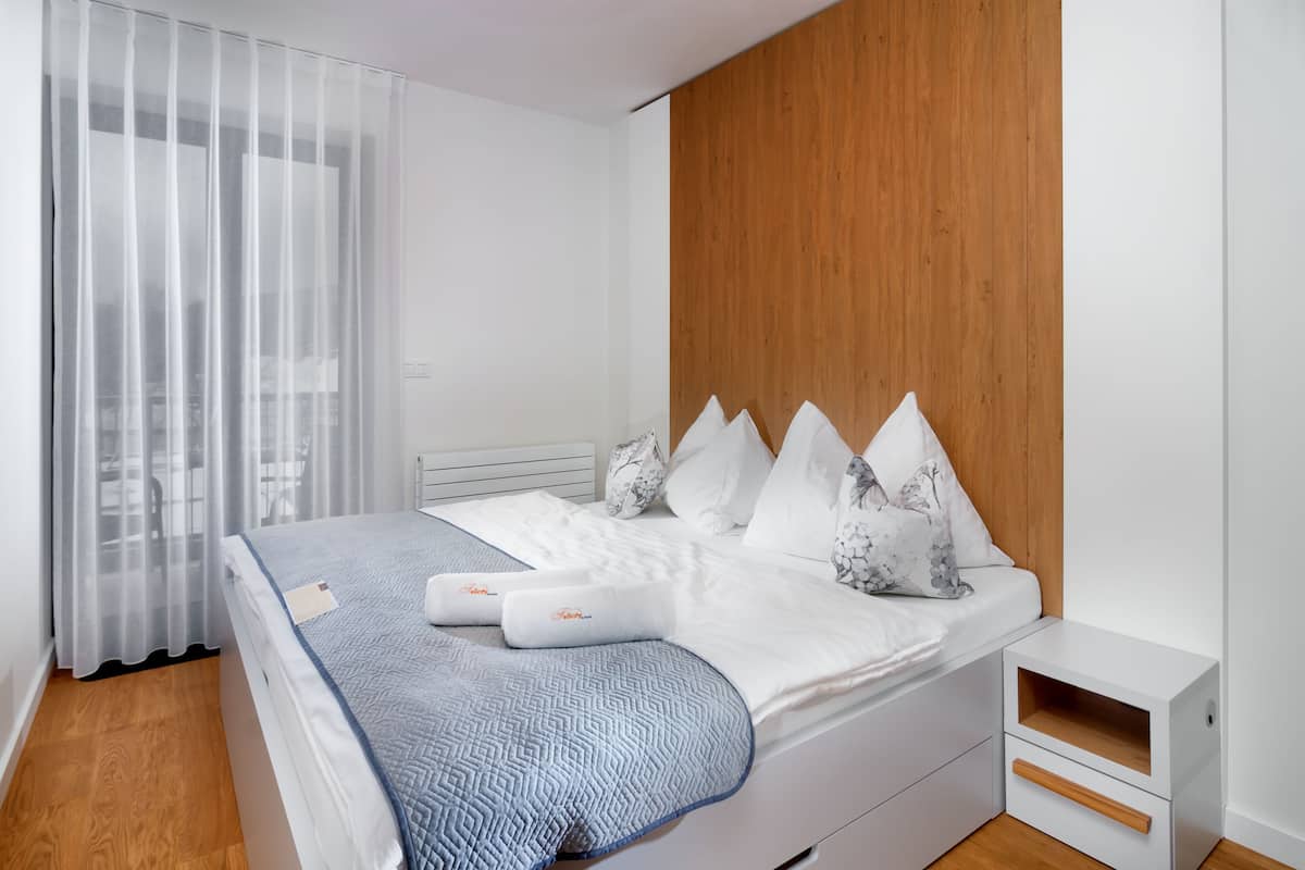 A modern bedroom with a bed, white bedding, a wooden headboard, a nightstand, and a window with sheer curtains.