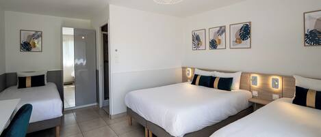A hotel room with three beds, a desk, and a chair. There are wall-mounted lights and framed artwork on the walls.