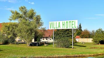 A sign for the Villa Hotel in a lush, green setting with a pond and picnic tables.
