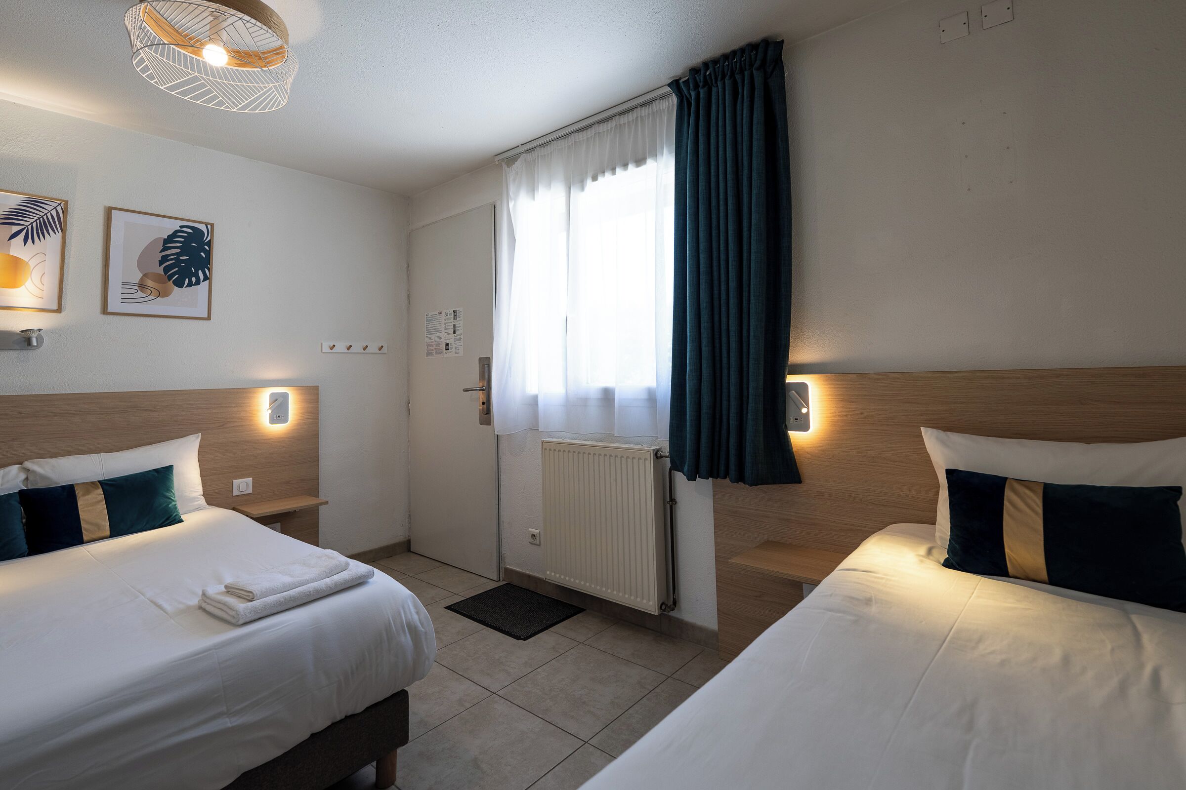 A hotel room with two beds, a window with curtains, wall-mounted lights, and artwork.