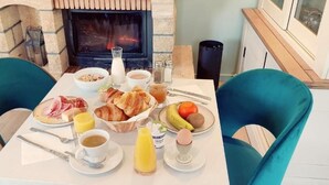 Mesa de pequeno-almoço posta com croissants, frutas e bebidas, com uma lareira ao fundo.