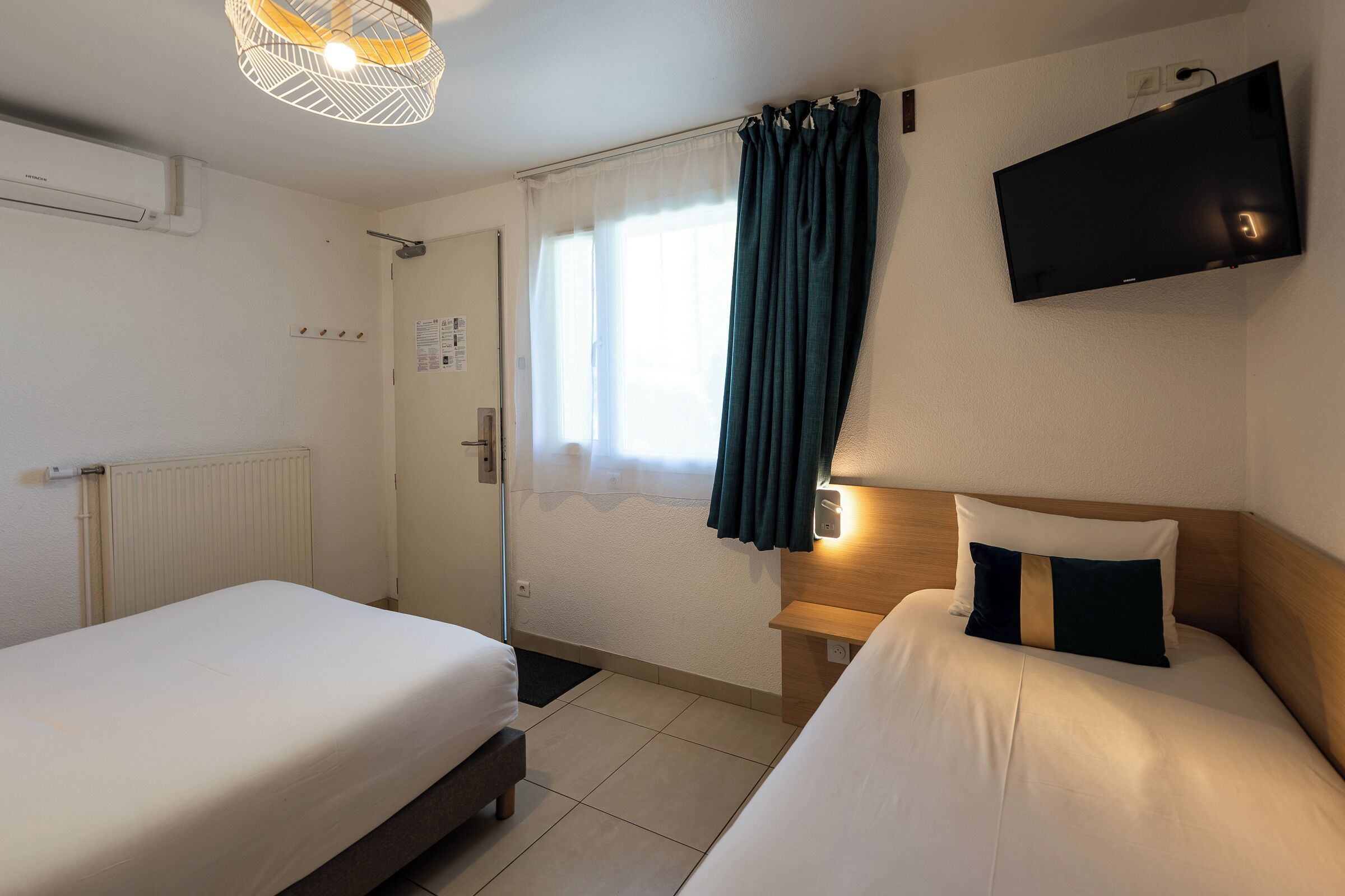 A hotel room with two beds, a TV, a window with curtains, and a radiator.