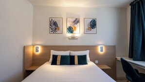 Uma cama bem arrumada com duas almofadas, uma mesinha de cabeceira, uma cadeira e três quadros com folhas na parede.
