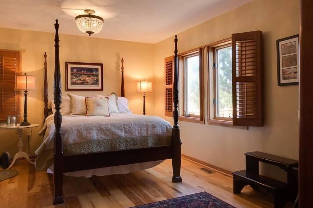 A bedroom with a four-poster bed, wooden flooring, a chandelier, and a large window with shutters.