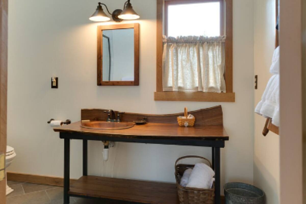A bathroom with a wooden vanity, a sink, a mirror, a window with curtains, a trash can, and a basket with towels.