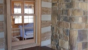 A rustic log cabin interior with a wooden desk, stone wall, and a window with a view of a blue chair and a lamp. - River Bluff Farm Bed & Breakfast (New Market)