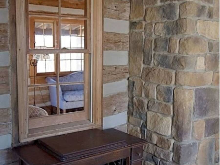 A rustic log cabin interior with a wooden desk, stone wall, and a window with a view of a blue chair and a lamp.