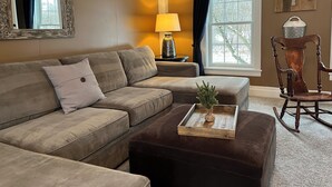 A living room with a sectional sofa, a small ottoman, a wooden rocking chair, a lamp, and a window with curtains. - Red Fox Run B&B (Cazenovia)