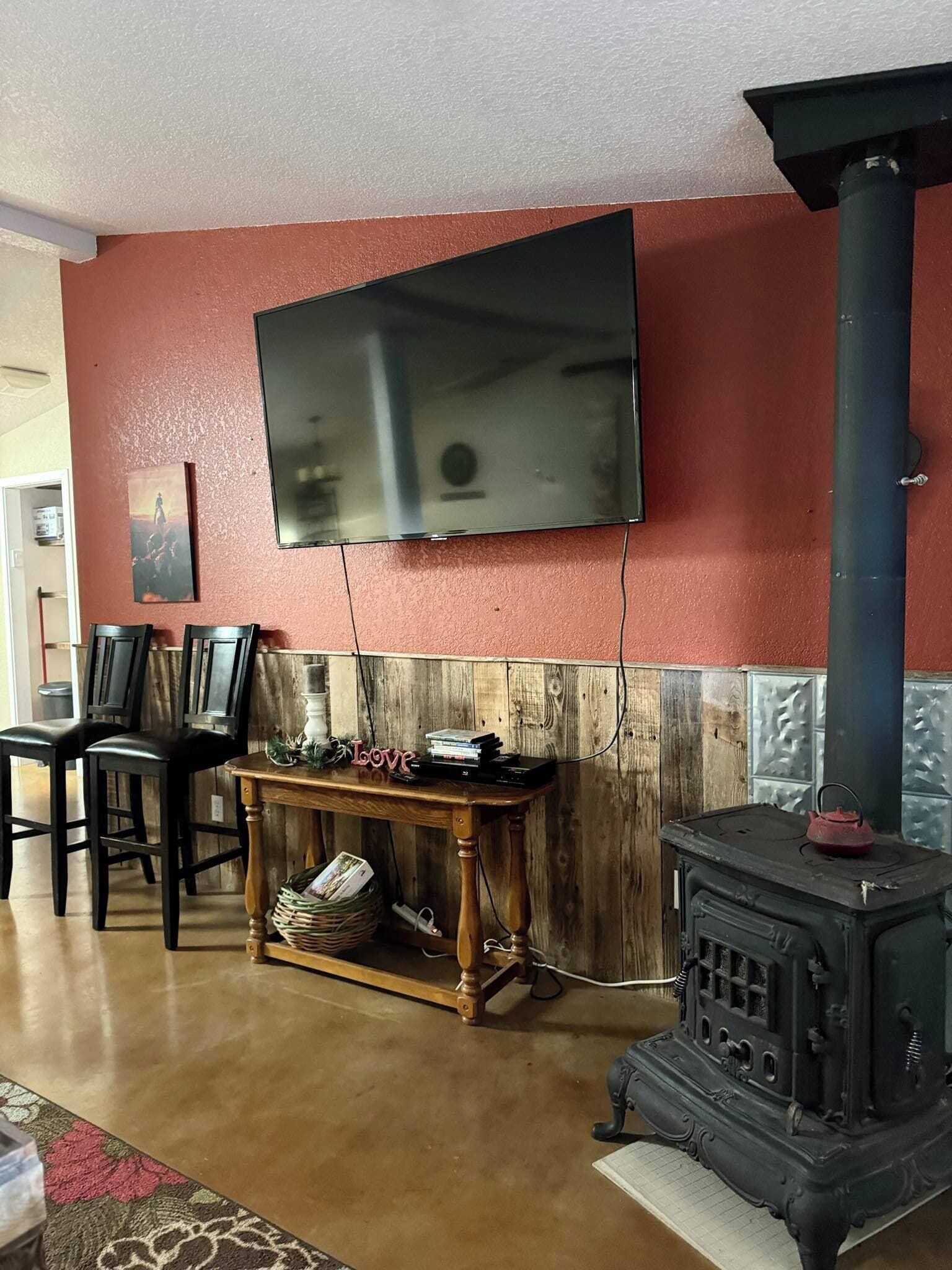 A living room with a TV mounted on a red wall, a wooden table with books, two chairs, and a vintage stove.