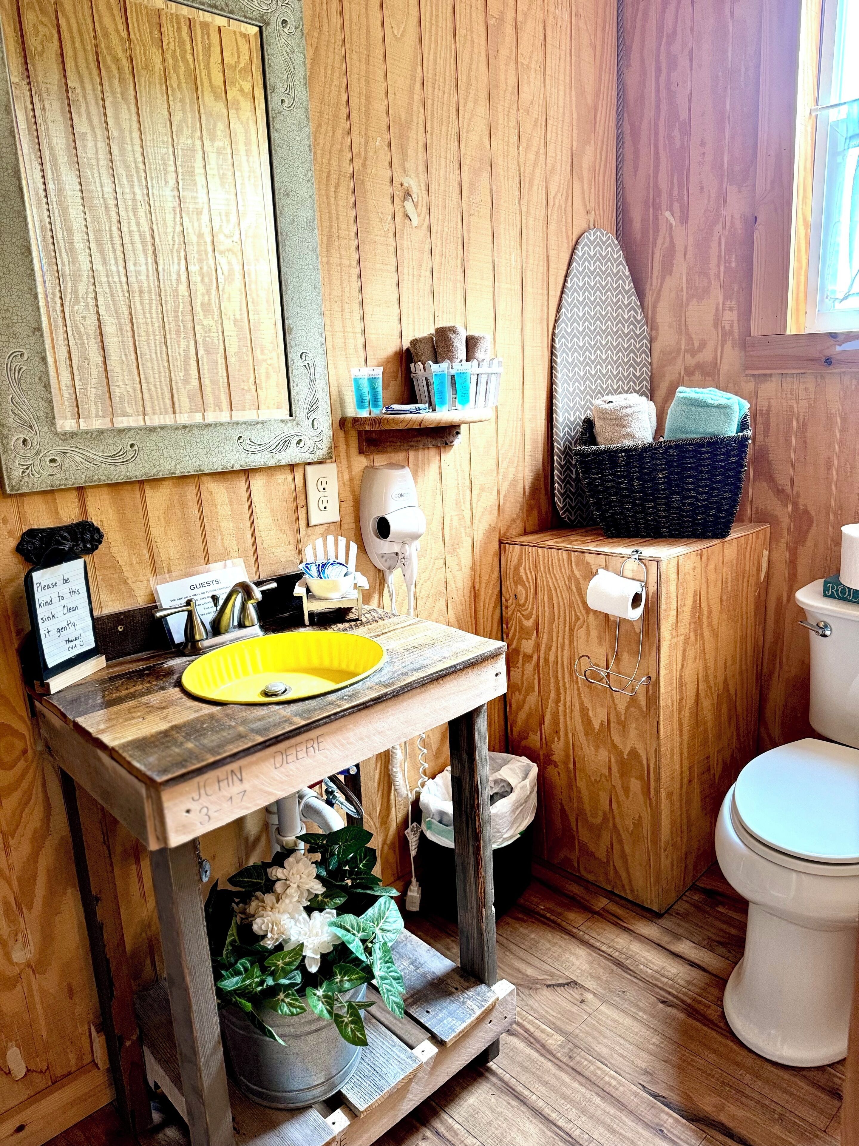 Ein rustikales Badezimmer mit einem Holzbadezimmer-Möbel, einem gelben Waschbecken, einer Toilette und einem Regal mit Pflegeprodukten.