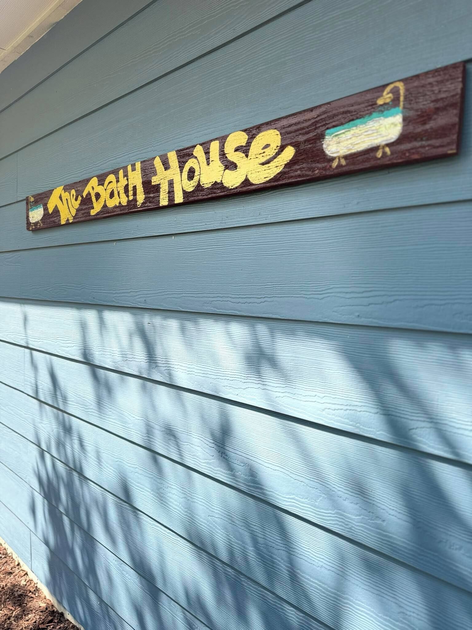 A wooden sign with the text "The Bath House" on a light blue house exterior.