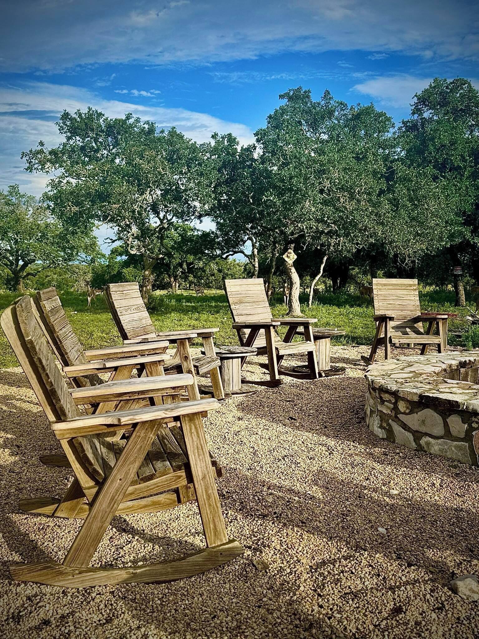 Outdoor wooden furniture arranged on a gravel surface with a stone fire pit, surrounded by trees and a clear sky.