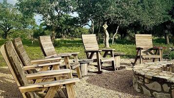 Outdoor wooden furniture arranged on a gravel surface with a stone fire pit, surrounded by trees and a clear sky.