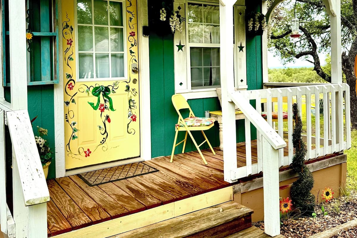 A cottage with a green exterior, a yellow door with floral decorations, and a small porch with a chair.