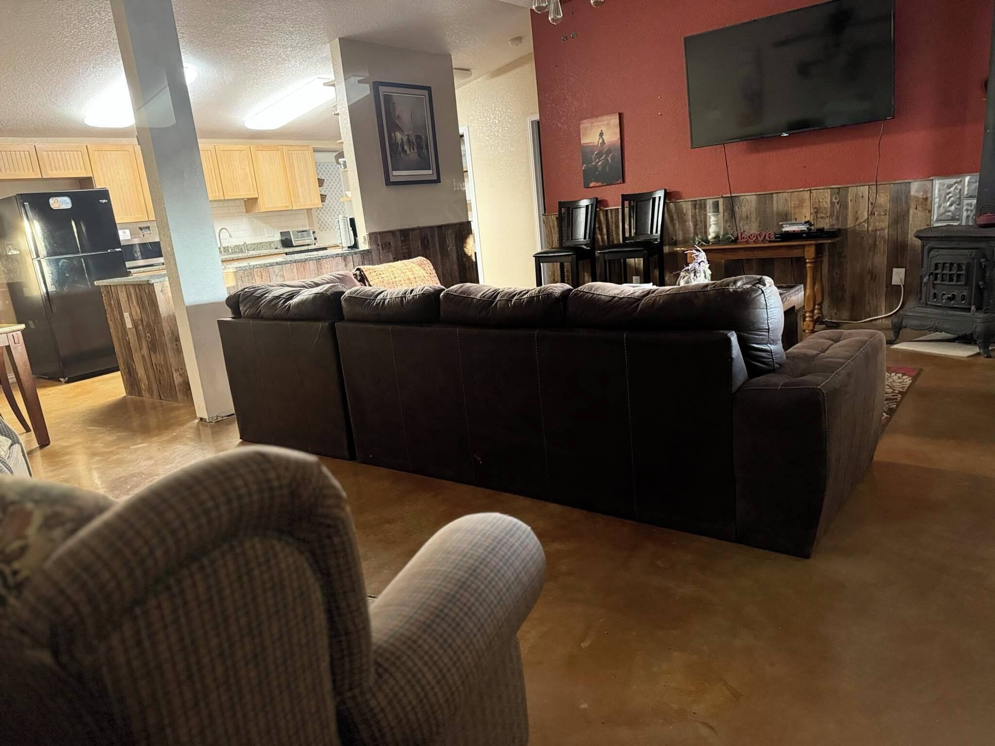A living room with a large sectional sofa, a TV mounted on the wall, and a kitchen area in the background.