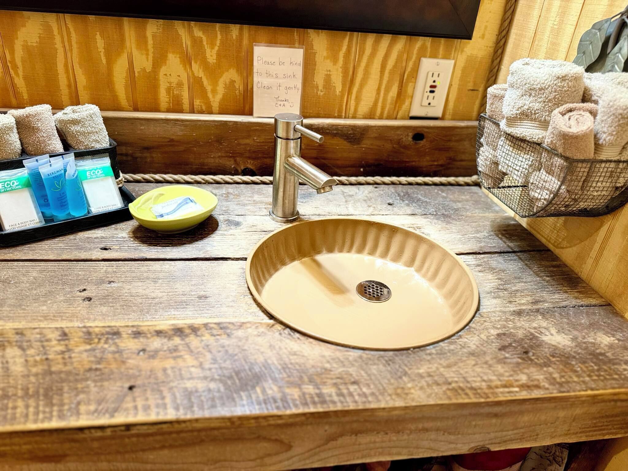 A wooden sink with a beige basin, a metal faucet, and a note asking to be kind to the sink. There are towels and a small basket of toiletries on the right.
