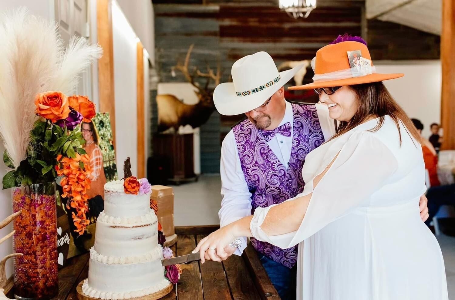 Un couple est en train de découper un gâteau blanc décoré de motifs floraux, sur une table en bois, dans un cadre rustique.