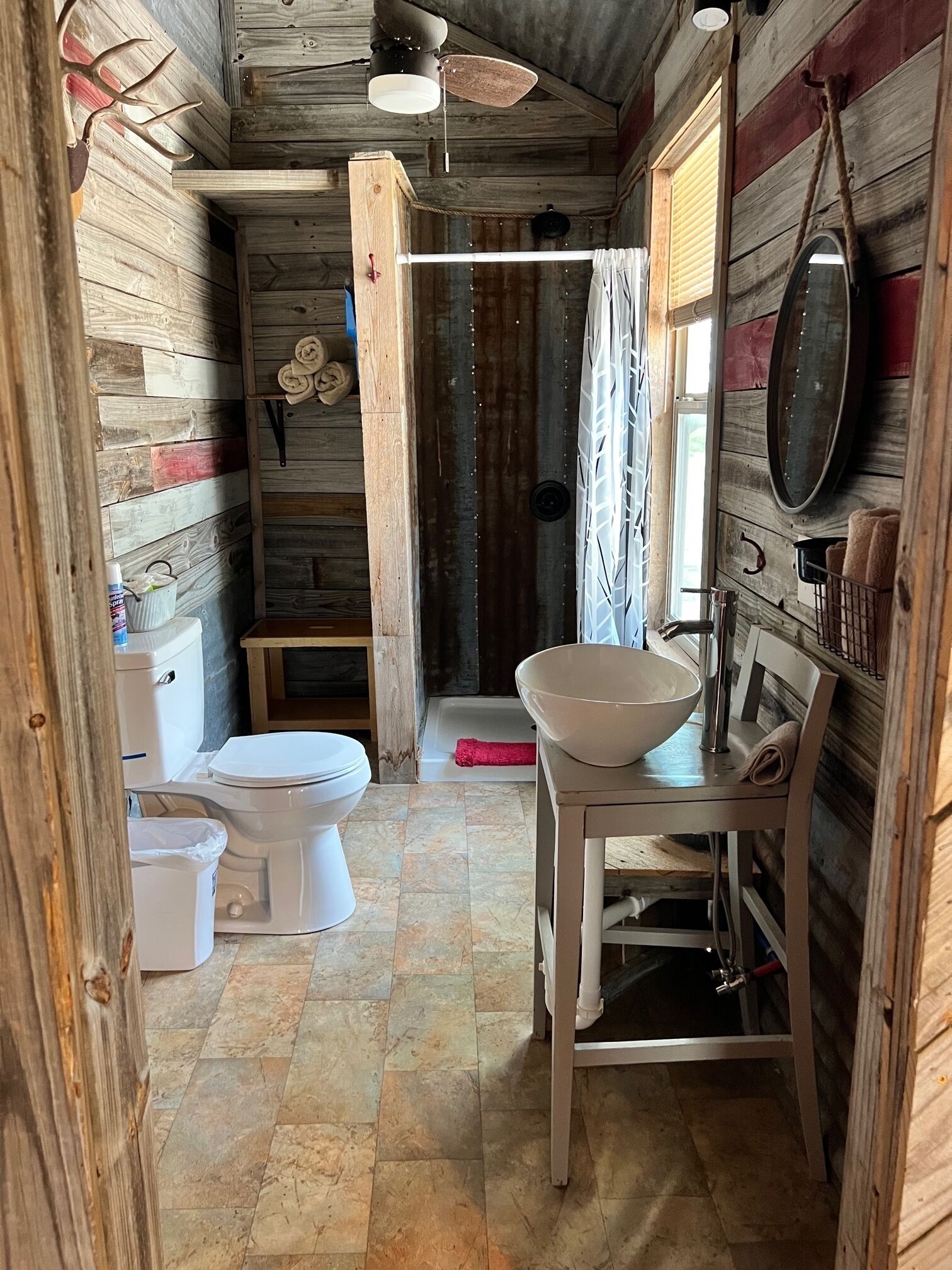 A rustic bathroom with wooden walls, a white toilet, a sink with a basin, and a ceiling fan.